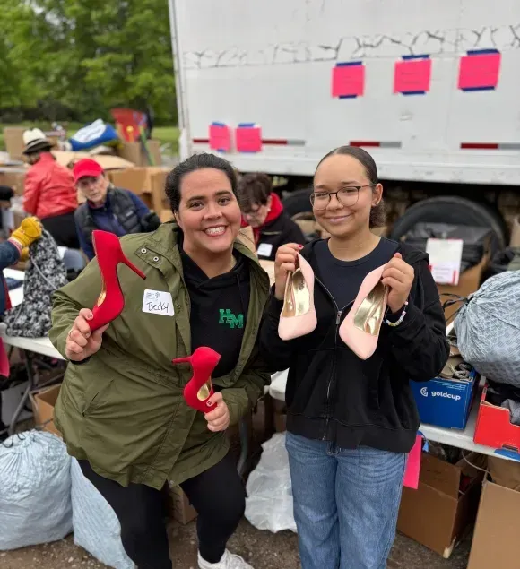 2 volunteers hold up shoes and smile as they help prepare for the North Oaks II Rummage Sale