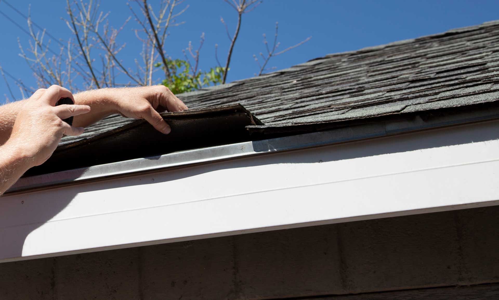 Hands lifting asphalt shingle on a roof. Blue sky in background.