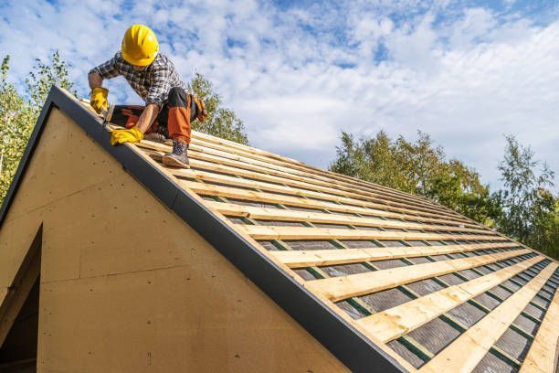 Roofer in yellow helmet installing roof shingles on a wooden frame outdoors under a blue sky.