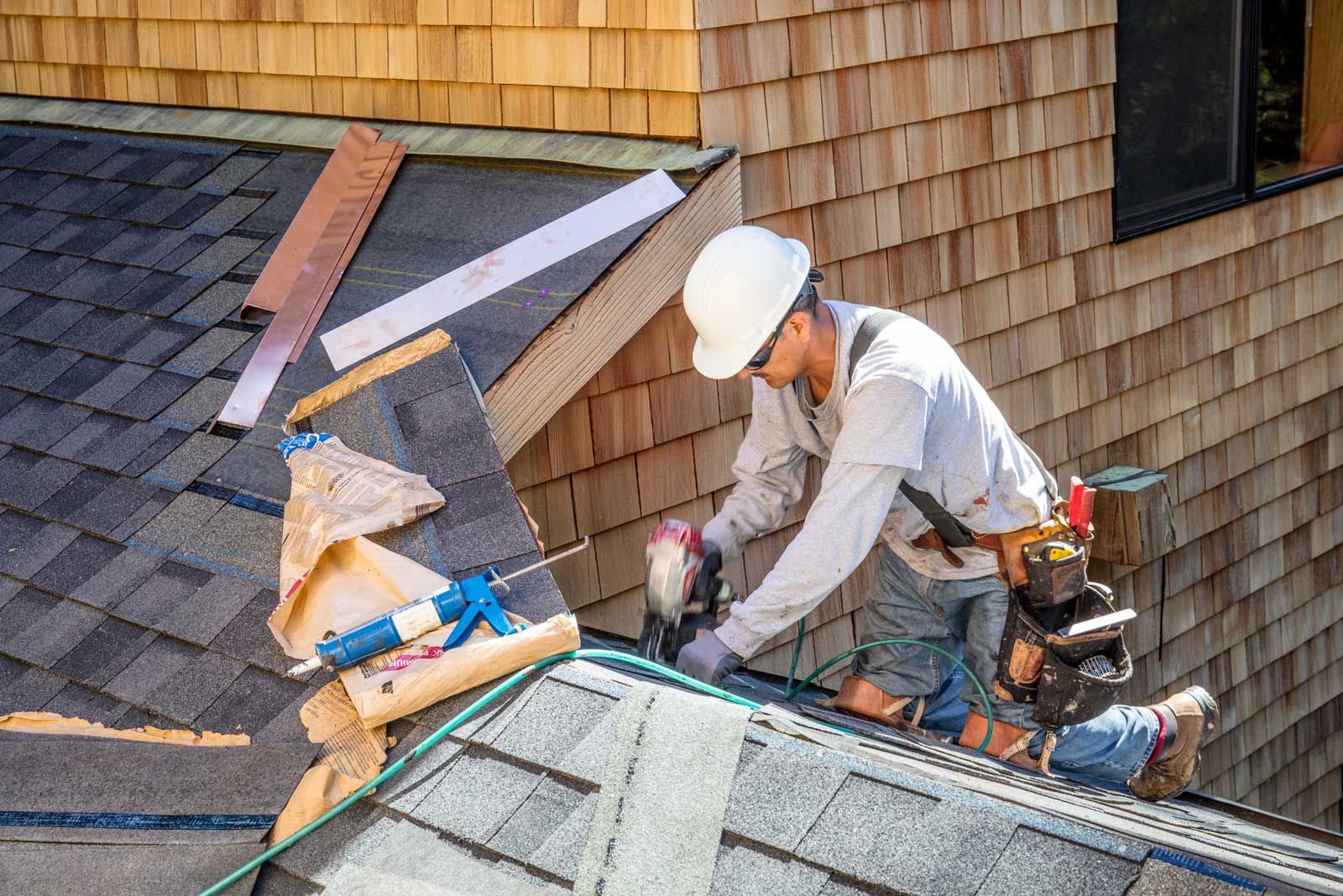 Roofer in hard hat using a nail gun on a shingle roof. Building materials and tools are visible.
