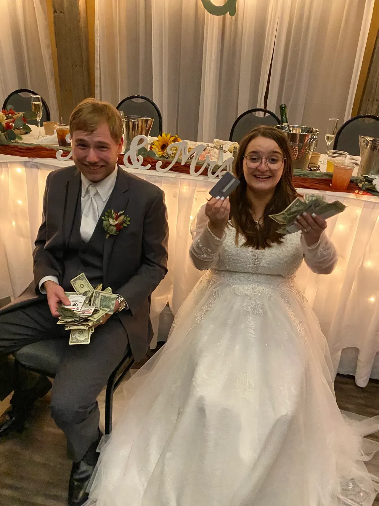 Newlyweds holding cash and cards at a wedding reception table decorated with flowers.