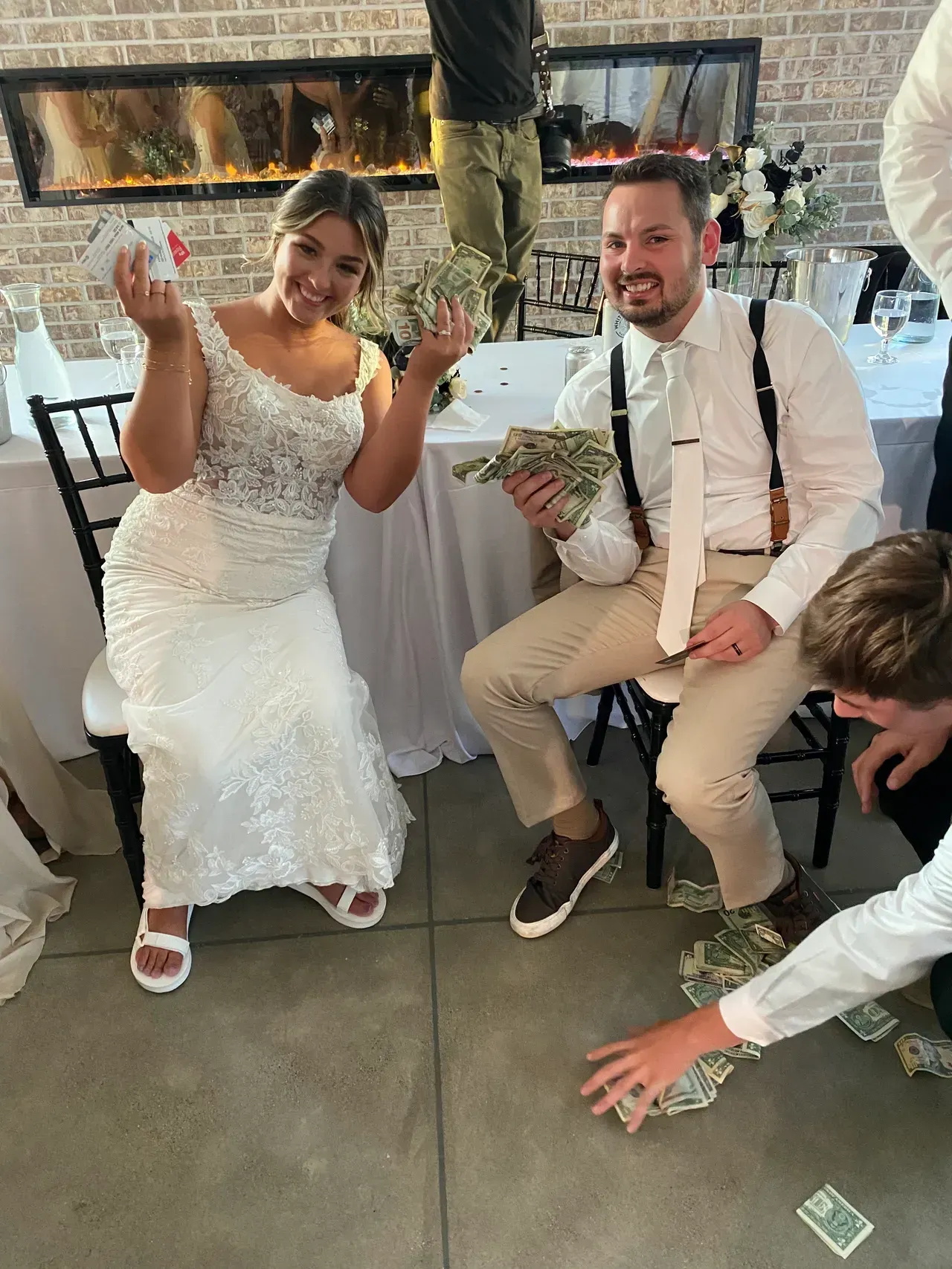 Bride and groom seated, holding money and herbs. People on the floor gather money. Wedding reception.