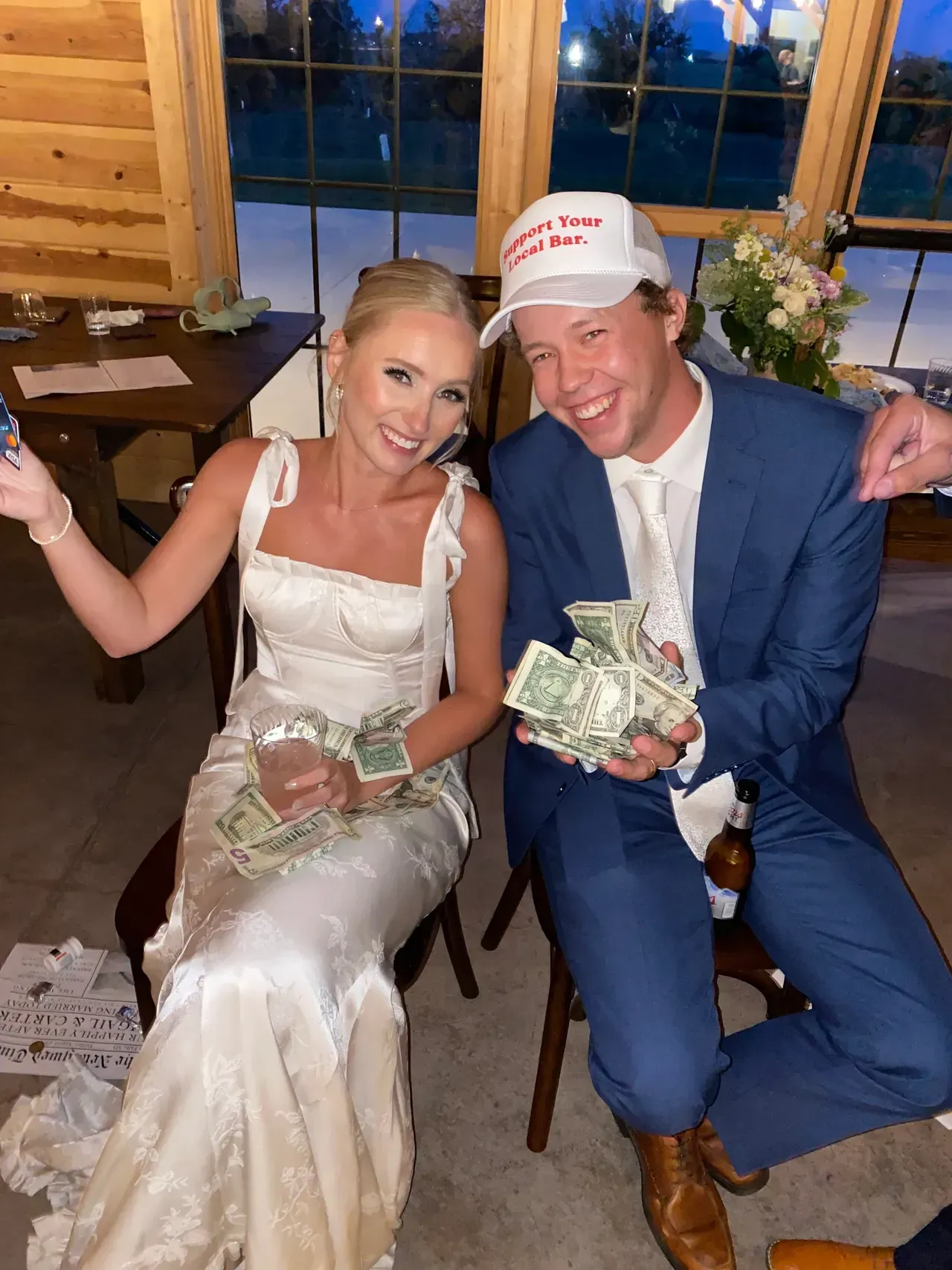 Bride and groom hold cash; she wears a white gown, he wears a blue suit and a white hat. Both are smiling.