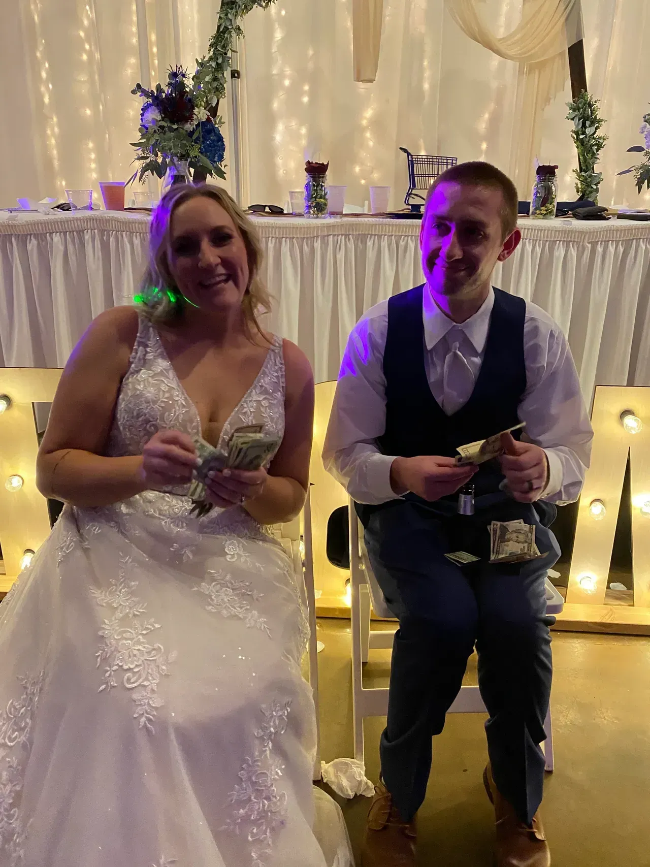 Bride and groom seated, holding money during a wedding reception. They smile at the camera. Lit backdrop.