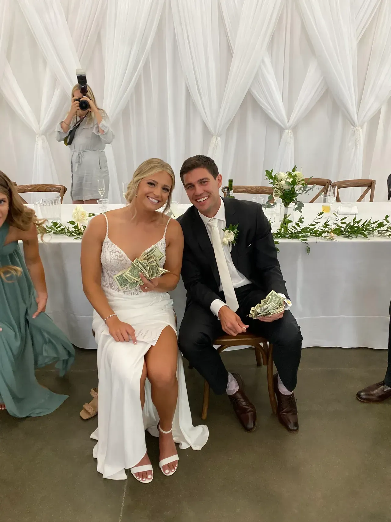 Bride and groom seated, smiling, holding cash, at a wedding reception. White dress, black suit, floral backdrop.