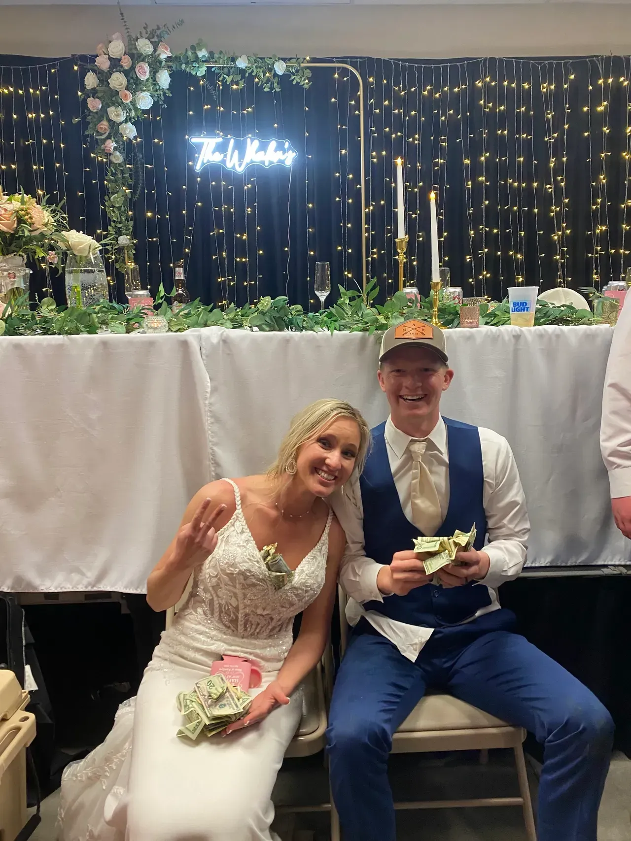 Bride and groom holding cash at wedding reception, smiles. White dress, blue suit, decorated table, backdrop.