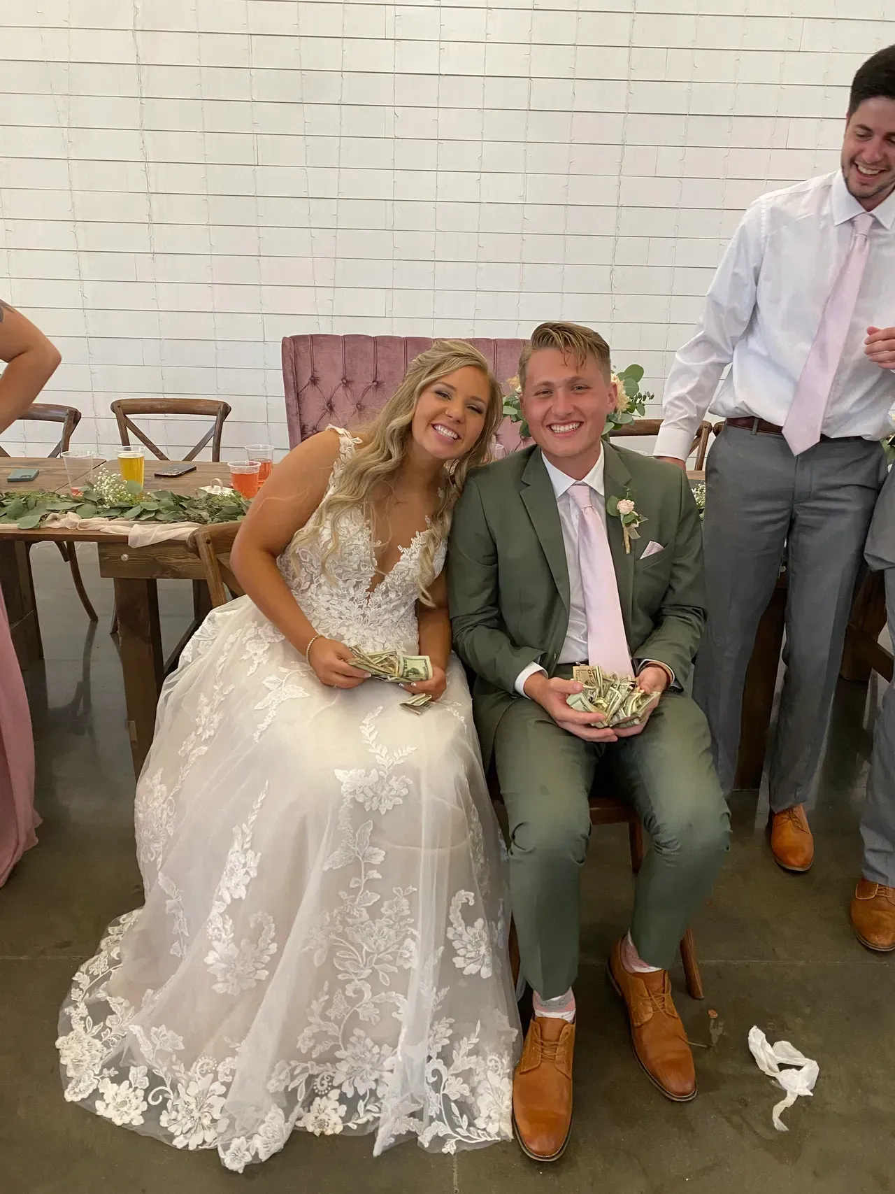 Bride and groom seated, smiling, holding small flowers; man in background laughs. Wedding reception.