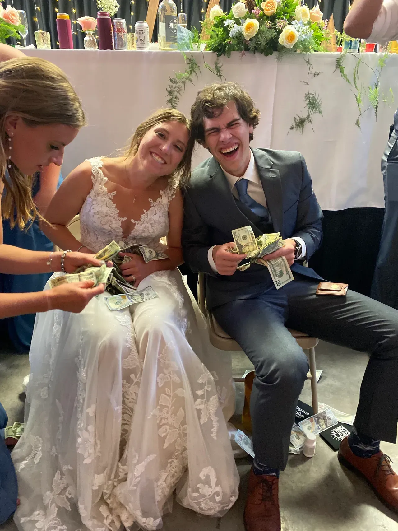 Bride and groom smiling, holding cash, with a bridesmaid. Wedding reception setting.