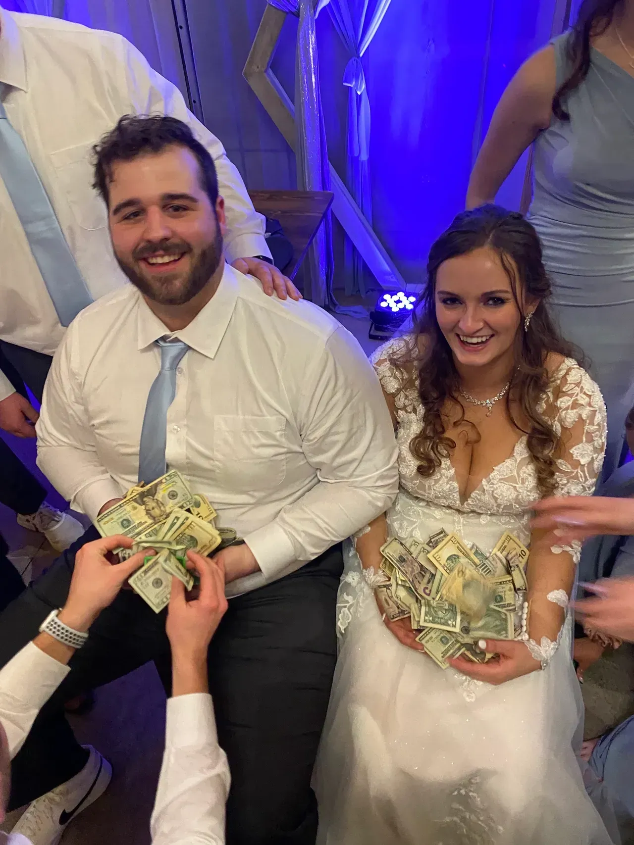 Wedding couple holds cash, smiling. People give money. Bride wears white dress; groom in a white shirt and blue tie.