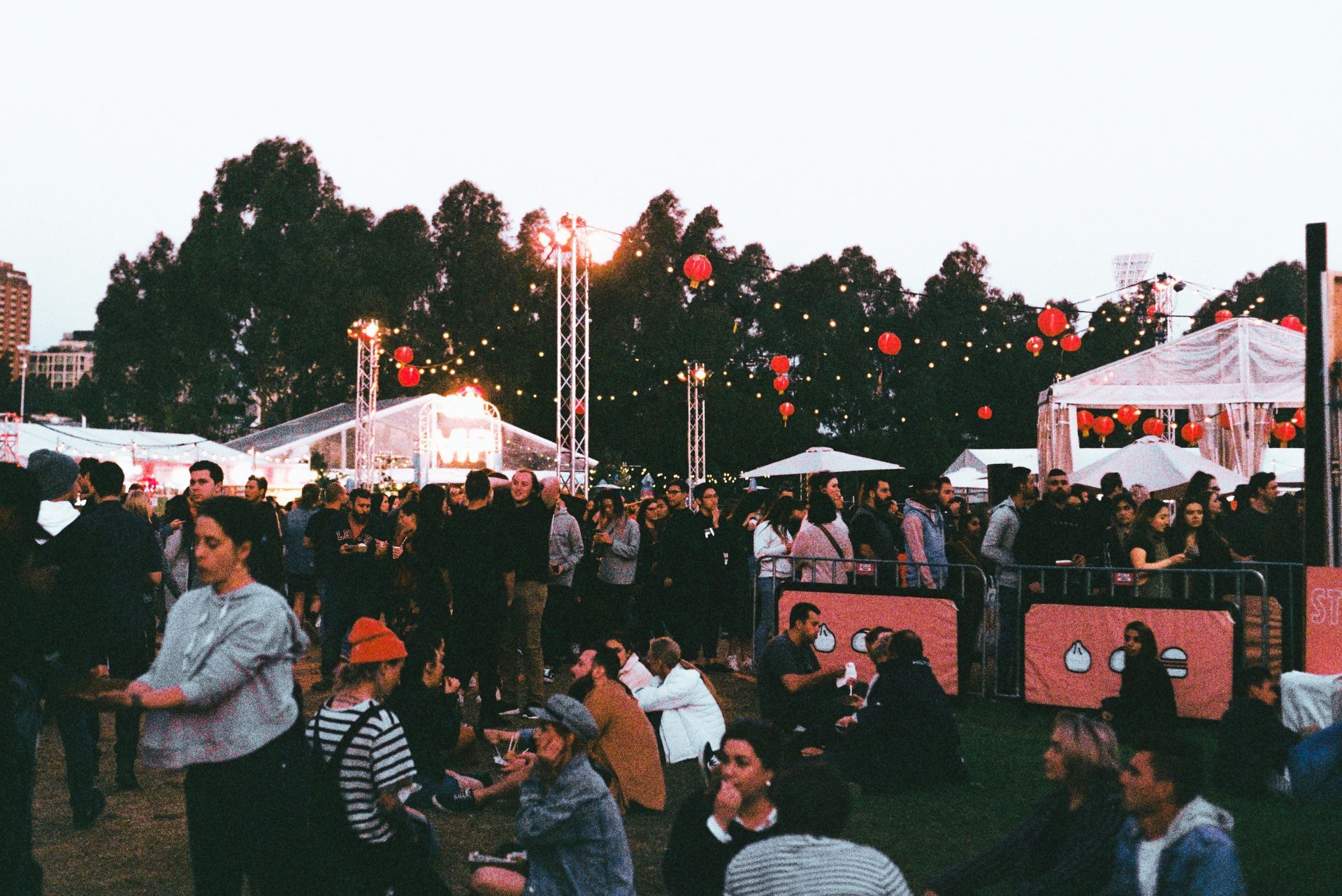 Large outdoor gathering with people; tents, lights, and trees in the background. Dusk setting.