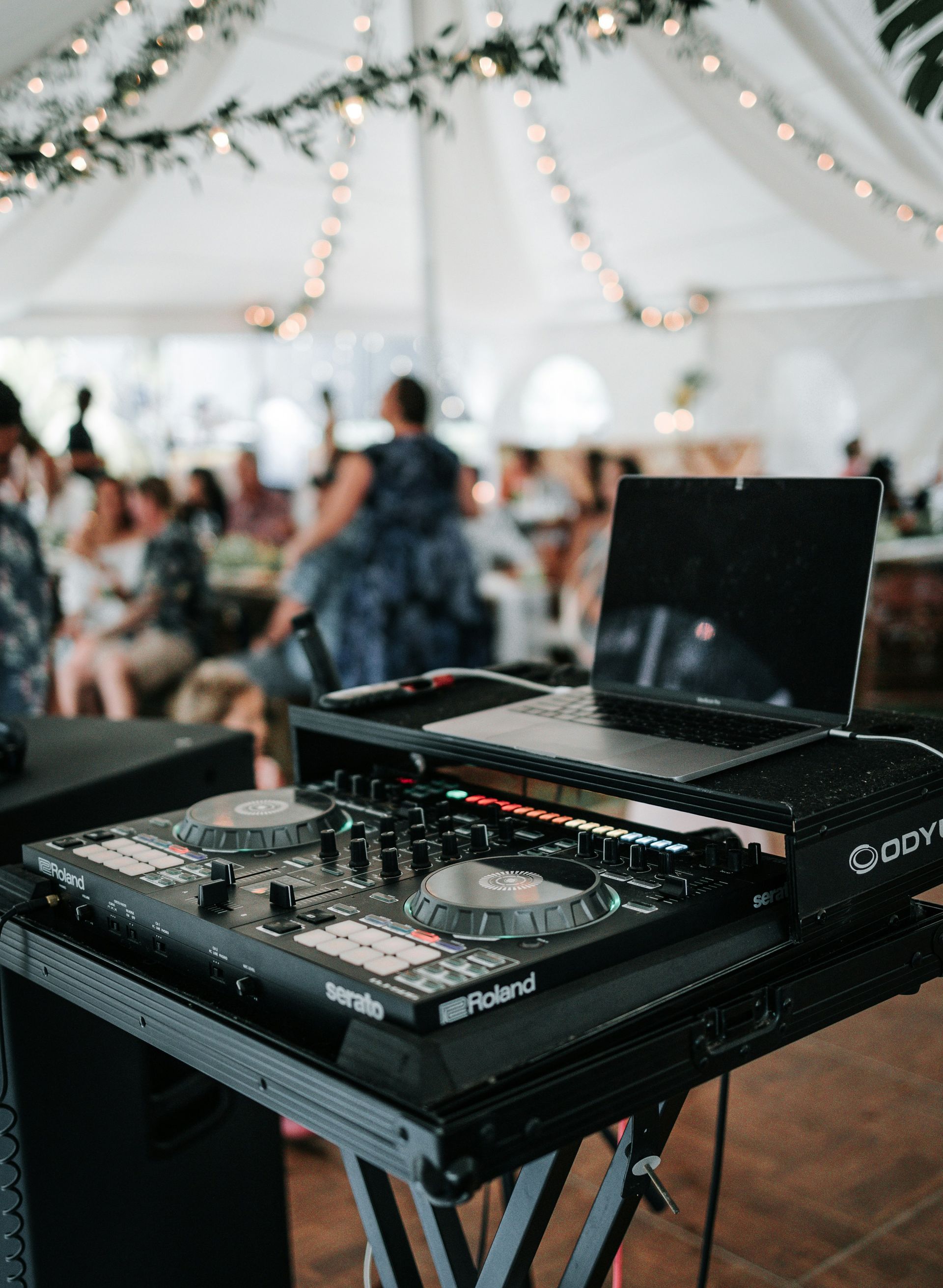 DJ setup at an event, with a laptop, mixer, and speakers under a tent. People in the blurred background.