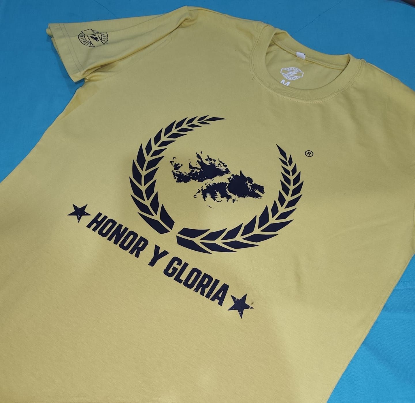Camiseta amarilla con diseño azul marino: una corona de laurel rodeando una silueta y la palabra 