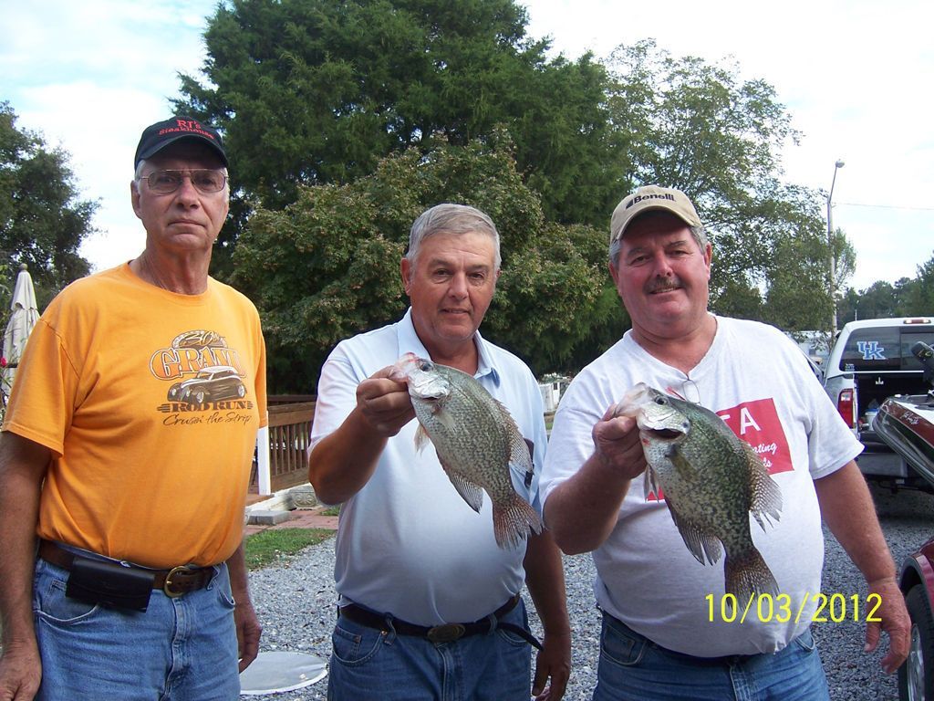 Three people standing outdoors in casual clothing; the two on the right hold up freshly caught fish.
