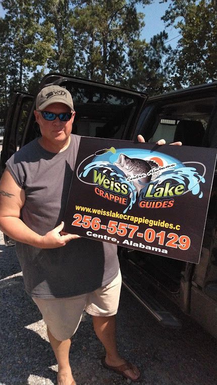 A person holding a Weiss Lake Crappie Guides sign with a phone number and location in front of a vehicle.
