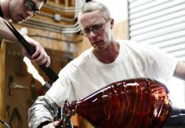Two people in a studio shape a glowing, textured red glass vessel at the end of a long metal rod.