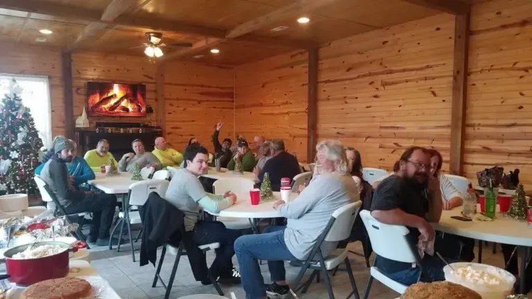 People sit at folding tables in a room with wood-paneled walls and a fireplace, gathered for a casual indoor event.