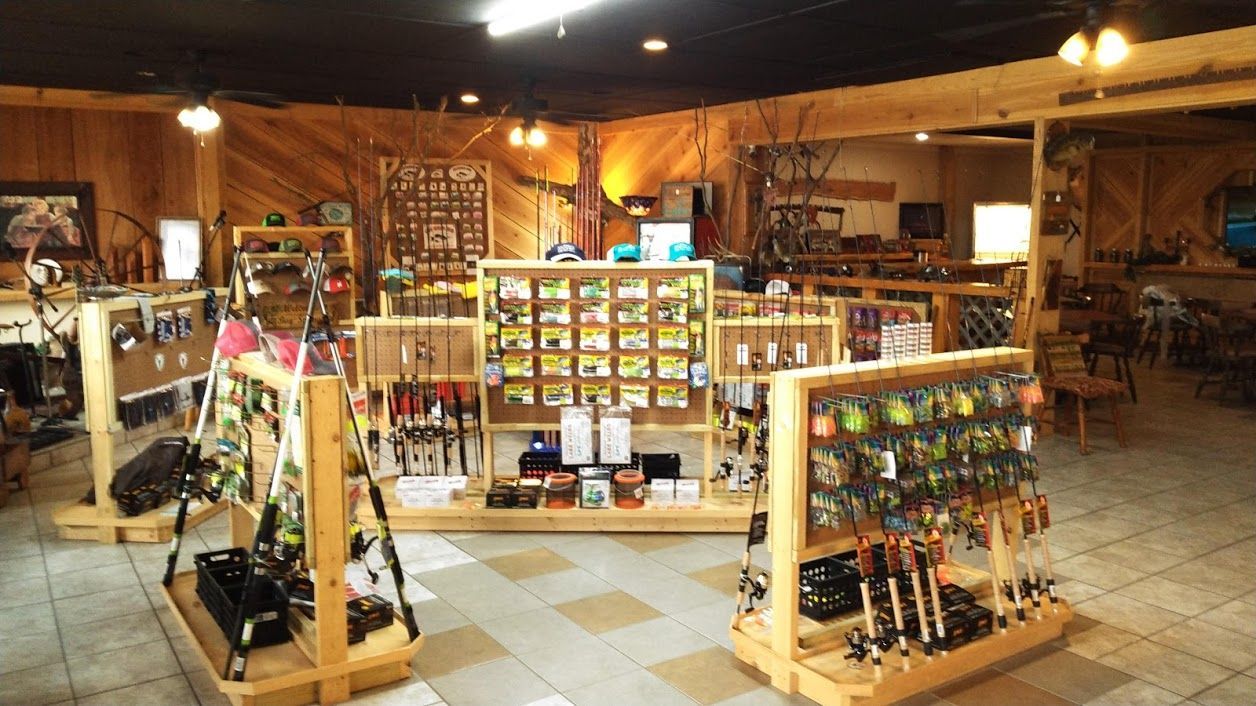 A retail store interior features wooden display stands filled with fishing equipment and gear under warm overhead lighting.
