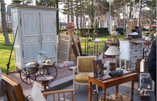 An outdoor antique market with a large pale blue cabinet, a vintage rocking horse, wooden chairs, and tables of items.