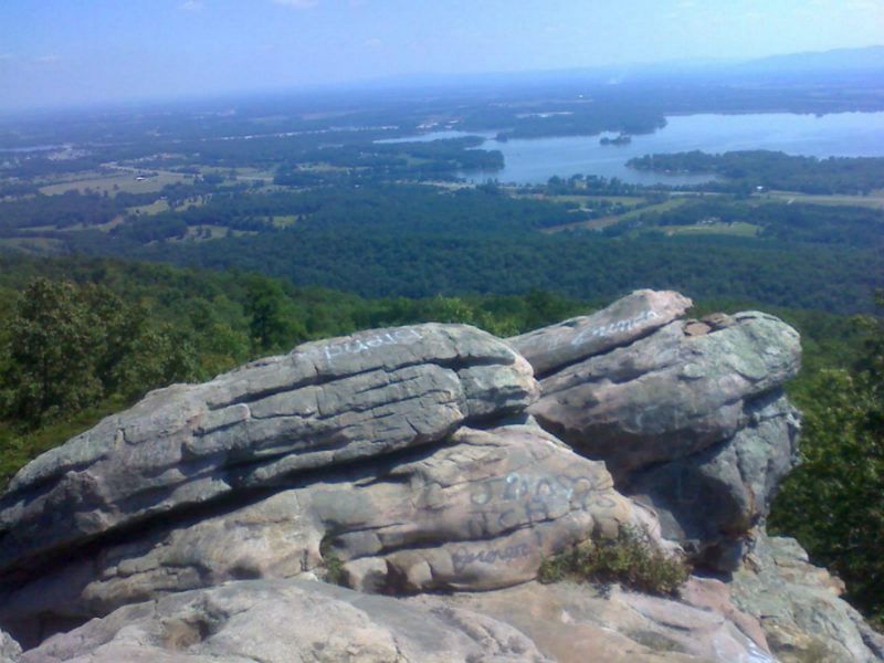 A rocky mountain summit overlooks a dense green forest and a large, distant lake under a clear blue sky.