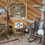 A wooden room with a fishing display on the wall, a fish artwork on a stand, and a rustic wooden rocking chair.