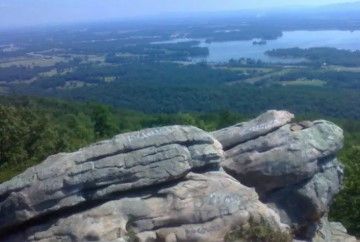 Large grey rock formations in the foreground overlook a vast valley with a lake and lush green forests.