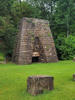 A historic stone blast furnace stands in a grassy clearing surrounded by lush green trees.