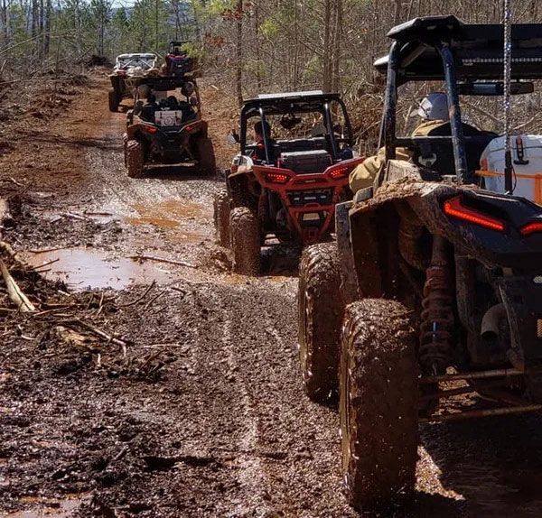 A line of side-by-side off-road vehicles drives through a muddy, forested trail.