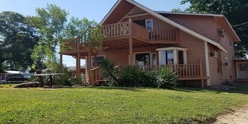 A two-story, light brown wooden house with a large deck, balcony, and a green lawn under a clear blue sky.