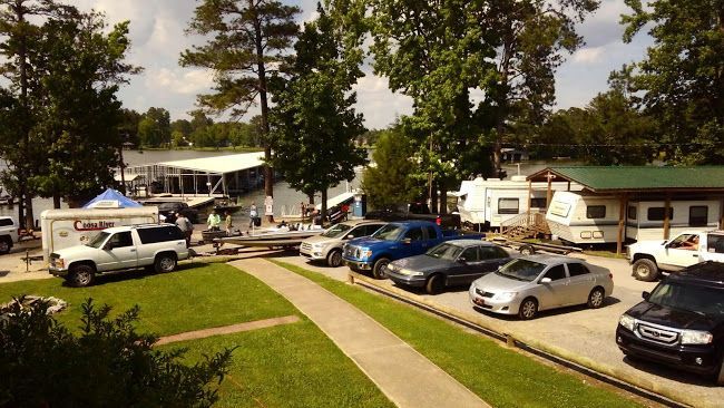 A sunny outdoor scene featuring a boat ramp, a parked car lot, and several campers near a tree-lined lakefront.