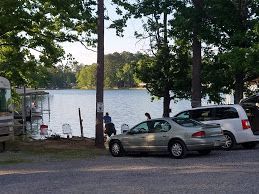 A silver sedan and a white minivan parked on a gravel lot overlooking a sunny lake with trees and people fishing.