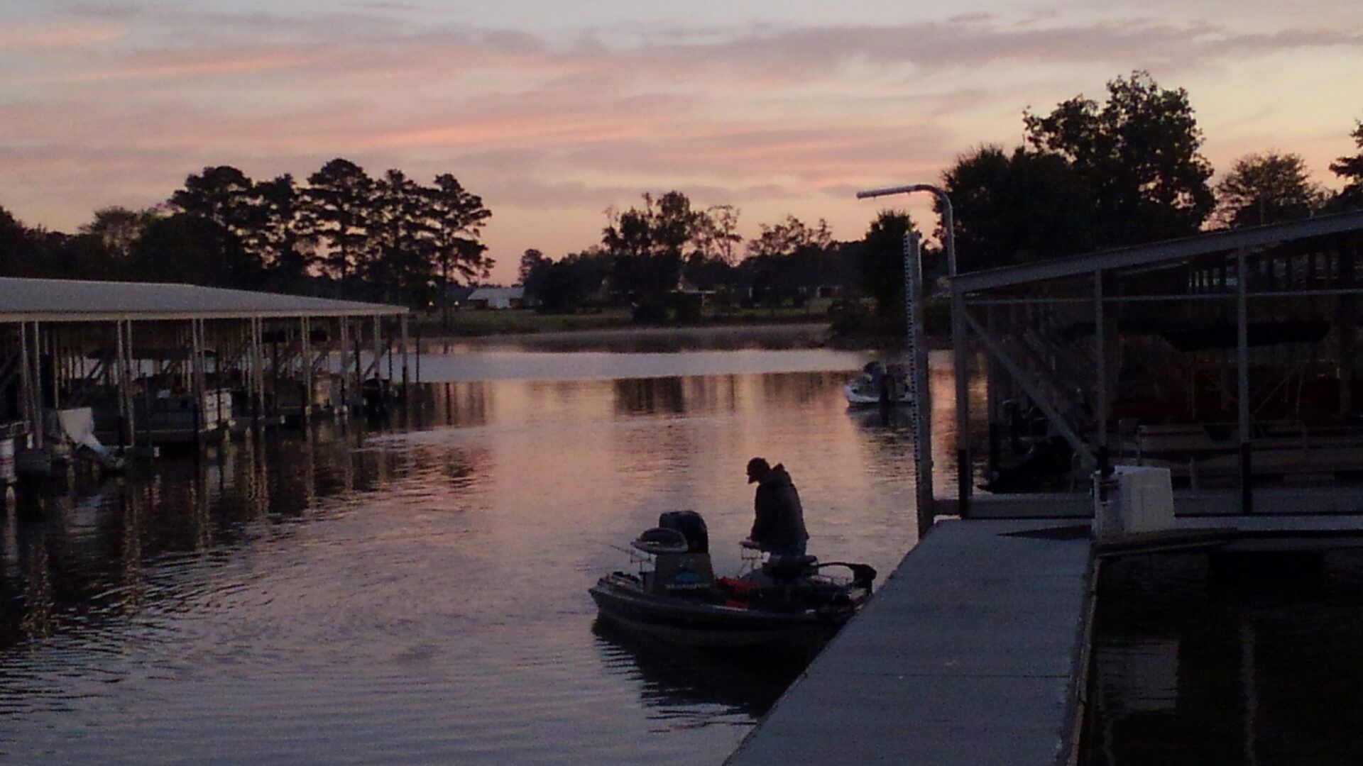 A person on a small fishing boat at a marina during a sunset with a pink and orange sky.