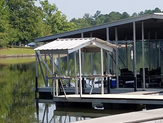 A small, white-roofed cleaning station sits on a wooden dock next to a large, metal-roofed boat slip on a calm lake.