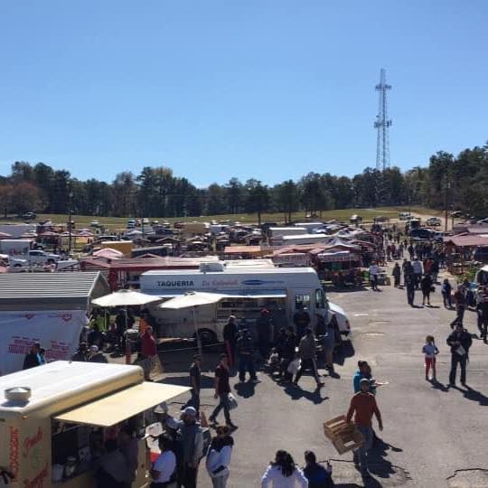 People walk through a sunny outdoor flea market filled with rows of vendor tents and food trucks under a clear blue sky.