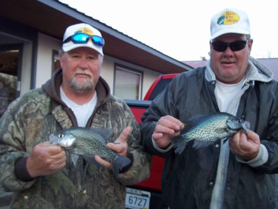 Two people stand outdoors wearing hats and holding freshly caught crappie fish in front of a red truck.