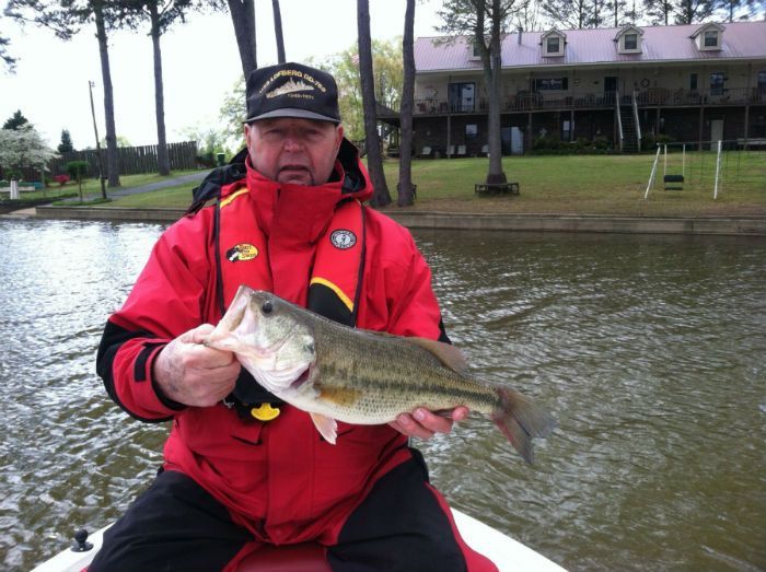 A person in a red fishing jacket holds a largemouth bass over the water in front of a house on a grassy bank.