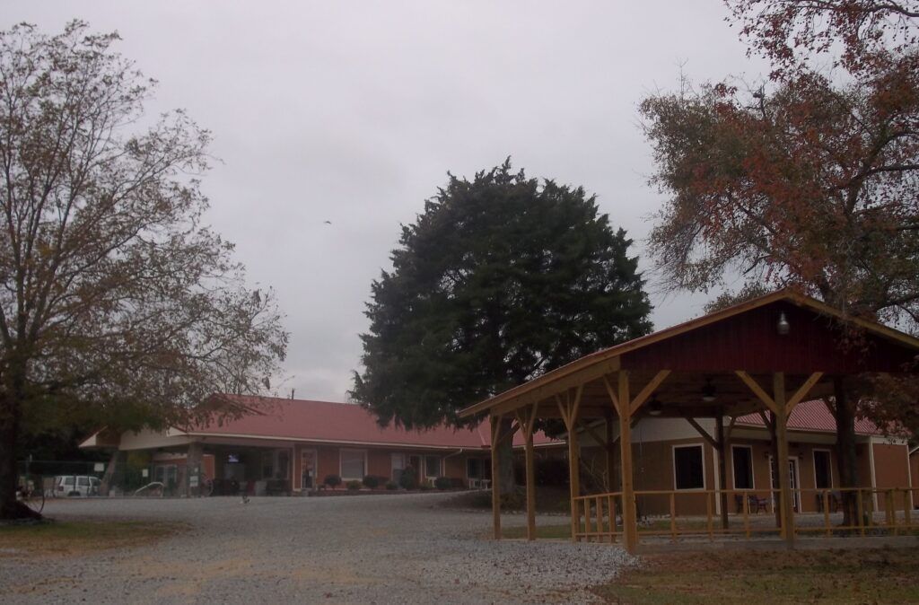 A gravel courtyard with a wooden pavilion on the right and a long, red-roofed building in the back under an overcast sky.
