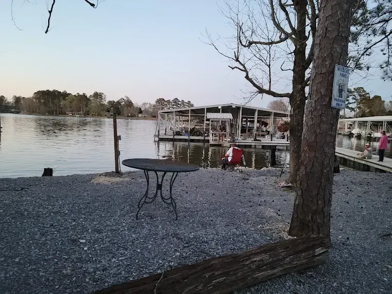 A gravel waterfront area with an outdoor table, looking out toward a boat dock on a calm lake at sunset.