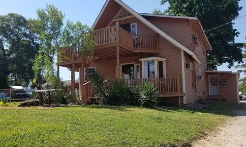 Two-story peach-colored house with a wooden balcony and wrap-around porch, set on a grassy lot under a clear blue sky.