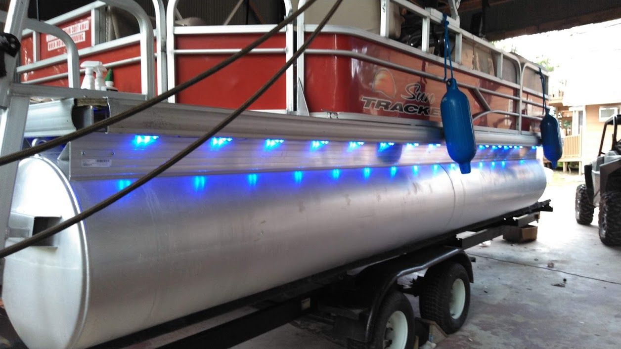 A pontoon boat sits on a trailer, featuring a decorative row of blue LED lights installed along the side of its hull.