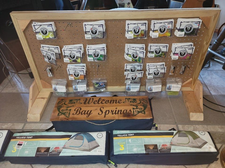 A pegboard display of small fishing lure packages sits above a welcome sign and two boxed camping tents on a floor.