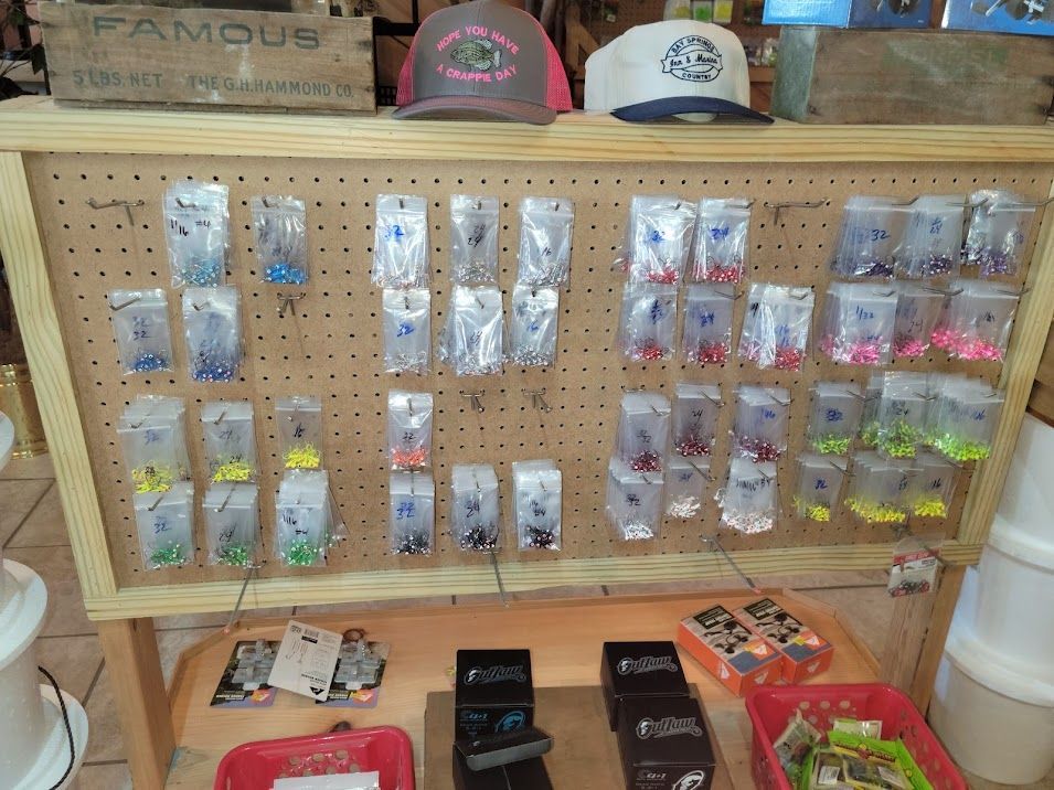 A pegboard display in a shop features many small, clear plastic bags of colorful fishing lures, with hats on top.