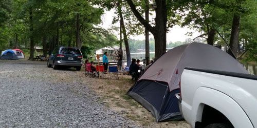 People camp near a lake with tents, cars, and chairs parked on a gravel ground under shade trees.
