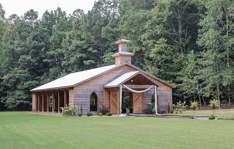 A rustic wooden chapel with a white metal roof and open front porch, set in a grassy clearing before a dense forest.