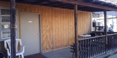 A small, wooden-paneled building with a porch, a door, a window, a white plastic chair, and outdoor patio furniture.