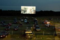 A drive-in movie theater at night with cars parked on a grass field facing a large glowing projection screen.