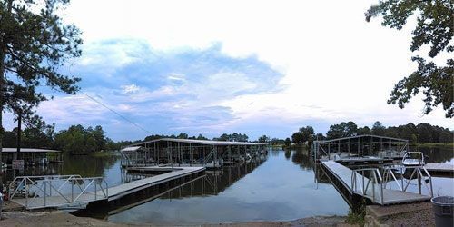 Floating boat docks on a calm lake under a cloudy sky, with metal ramps leading out from the grassy shoreline.