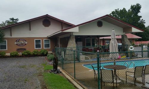 A tan and white motel building with a gabled roof and stone pillars, featuring a fenced-in swimming pool in the foreground.