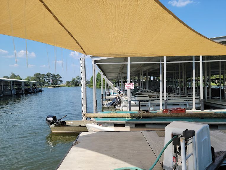 A tan shade sail covers boat slips at a marina, with a small flat-bottomed boat moored nearby on a sunny day.