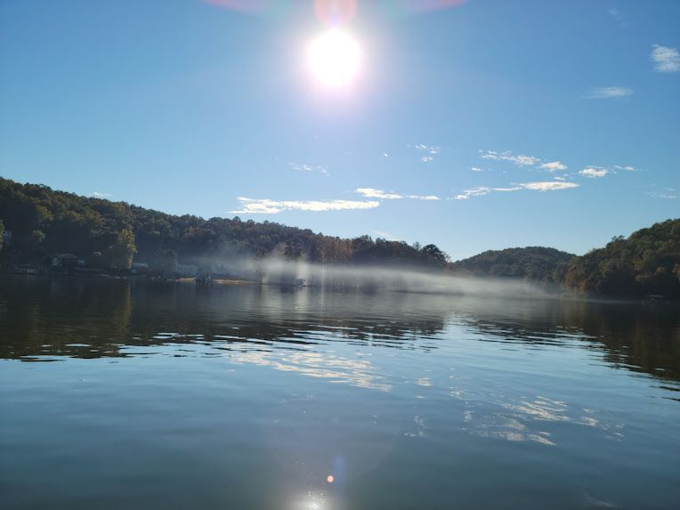 Sunlight shines over a calm, blue lake with a low layer of morning fog drifting above the water near tree-covered hills.