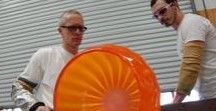 Two glassblowers in protective gear work with a glowing, orange circular glass piece in a studio.